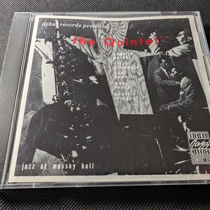 May include: A compact disc case featuring the album Jazz at Massey Hall by The Quintet. The cover art is a black and white collage of musicians including Charlie Parker, Dizzy Gillespie, Bud Powell, Max Roach, and Charlie Mingus, with the text debut records presents, the Quintet, and Original Jazz Classics Compact Disc.
