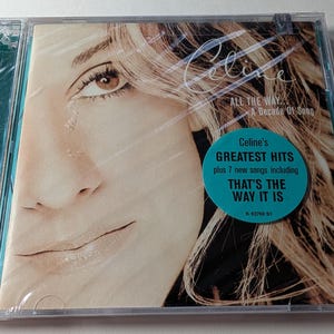Puede incluir: Una caja de CD con la foto de una mujer rubia. El CD se titula "All The Way... A Decade Of Song" de Celine. Una pegatina turquesa dice "Celine's Greatest Hits" y "That's The Way It Is."