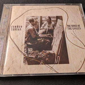 May include: A compact disc case featuring a sepia-toned photograph of two people working with fabric. The cover text reads Common Thread: The Songs of the Eagles. A thin, brown string is looped across the textured, light tan paper cover.