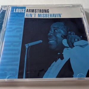 Puede incluir: Una caja de CD con el álbum de Louis Armstrong "Ain't Misbehavin'". La portada muestra una imagen en tonos azules de Armstrong cantando en un micrófono. La caja del CD es de plástico transparente.