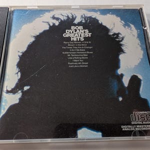 May include: A Bob Dylan's Greatest Hits CD in a clear plastic case. The album cover features a silhouette of a person with white accents against a blue background. The CD case includes a list of song titles and the Compact Disc logo.