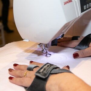 May include: A white sewing machine with a black and gray embroidery hoop. The machine is embroidering a white design on a white fabric. The person's hands are wearing black and gray finger cots.