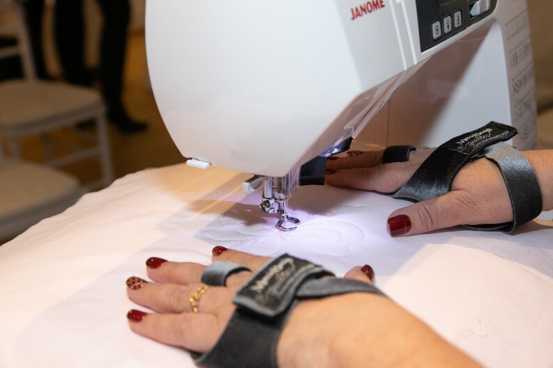 May include: A white sewing machine with a black and gray embroidery hoop. The machine is embroidering a white design on a white fabric. The person's hands are wearing black and gray finger cots.