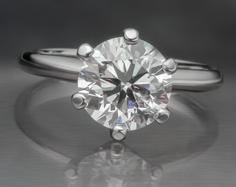 May include: A solitaire diamond ring with a round-cut diamond set in a polished silver band. The diamond is held in place by six prongs, showcasing its brilliance. The ring is displayed on a reflective surface.