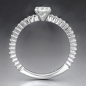 May include: A silver ring with a single, round diamond set in a prong setting. The band is decorated with a row of smaller diamonds.