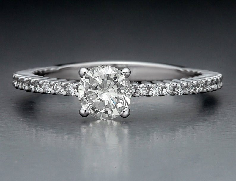 May include: A silver engagement ring with a round, clear center stone and a band of smaller, clear stones.