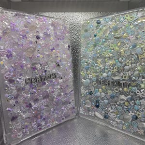 Peut inclure: Deux étuis en plastique transparent remplis de petites décorations colorées. Les étuis portent l'inscription "HARMONY" en paillettes argentées sur le devant.