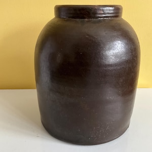 May include: A dark brown, glazed ceramic jar with a rounded body and a slightly narrower neck. The jar has a smooth, glossy finish and appears to be handmade. The jar is likely used for storage or decoration.