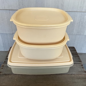 May include: A stack of three vintage Tupperware containers in shades of cream and off-white. The containers are square or rectangular with matching lids. The containers are stacked on top of each other.