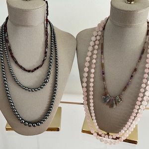 May include: Two necklaces displayed on mannequin busts. One features three strands of dark gray beads, while the other showcases three strands of pink beads. A third necklace has a mix of colorful beads.