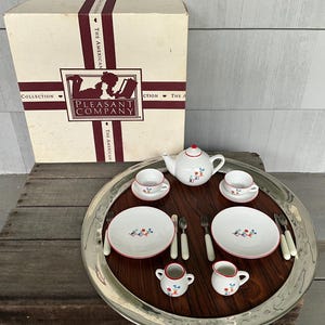 May include: A vintage children's tea set with a white teapot, cups, saucers, plates, and serving pieces, all with red trim and floral designs. The set is arranged on a silver tray with a wooden base, next to a box with the text "Pleasant Company".