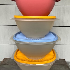 May include: A stack of four plastic mixing bowls with lids. The top bowl is coral, the next three are clear with yellow, blue, and yellow lids. The bowls are designed for food preparation and storage.