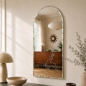 May include: An arched wall mirror with a scalloped edge and a white line drawing of a deer etched onto the glass surface. The mirror reflects a room interior featuring a wooden sideboard, a sunburst clock, and a pendant light fixture.