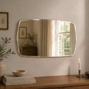 May include: A rectangular wall mirror with slightly rounded corners and a beveled edge. The mirror reflects a room interior with a window, a wooden cabinet, and a potted plant. The glass surface shows subtle signs of aging and patina.