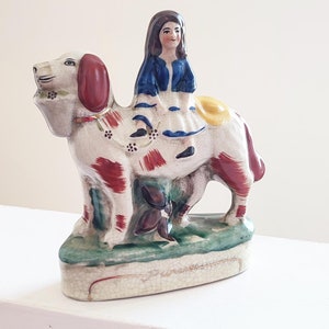 Puede incluir: Una figurita de cerámica de una niña con un vestido azul montando un perro blanco con marcas rojas y marrones. El perro está de pie sobre una base verde con flores pintadas.