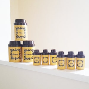 Può includere: Un set di dieci barattoli in ceramica gialli e marroni con scritte nere. I barattoli sono etichettati con "Tè", "Zucchero", "Caffè", "Timo", "Rosmarino", "Origano", "Cannella", "Prezzemolo" e "Salvia".