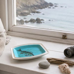 May include: A rectangular ceramic dish with a white exterior and a bright turquoise interior featuring a dark illustration of a fish. The dish is positioned on a white ledge alongside a smooth grey stone, a piece of driftwood, a seashell, and binoculars.
