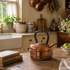 May include: A copper tea kettle with a black handle sits on a wooden table next to a stack of three books titled Cakes and Ale, Plain Cookery, and The English Kitchen. A white and blue patterned pitcher with flowers is nearby on the table.