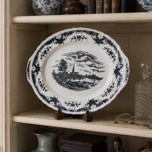 May include: An oval ceramic platter featuring a dark blue transferware landscape scene with a central church spire, rolling hills, and clouds. The rim is decorated with a repeating floral and scroll pattern on a white background, displayed on a stand.