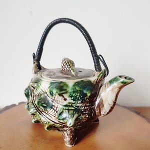 May include: A green and brown ceramic teapot with a black woven handle. The teapot has a textured, shell-like design and a small, round lid with a knob in the shape of a pine cone.