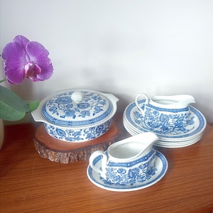 May include: A blue and white porcelain dinnerware set featuring a covered casserole dish, gravy boats, and plates. The floral pattern adorns each piece. The items are displayed on a wooden surface, with a purple orchid adding a touch of colour.