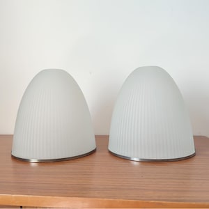 May include: Two frosted glass lampshades with a ribbed design. The lampshades have a rounded, bell-like shape and are trimmed with a silver-coloured metal band at the base. They sit on a wooden surface.