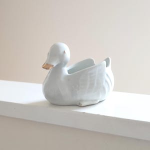 May include: White ceramic duck planter with a gold beak. The duck is lying on its side with its wings spread out.