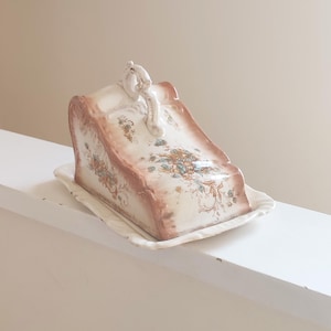 May include: A vintage porcelain cheese dish with a floral design. The dish has a lid with a scroll-shaped handle and a scalloped edge.