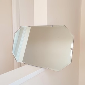 May include: An octagonal frameless mirror with beveled edges. The mirror is mounted with small, silver-colored clips. The glass reflects the surrounding light and colors, creating a simple, elegant aesthetic. The mirror is a decorative item.
