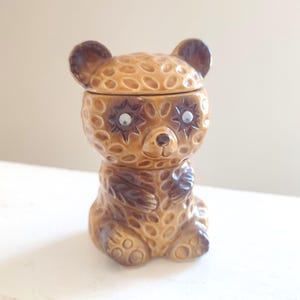 May include: A ceramic bear-shaped container with a lid. The bear is primarily brown with darker brown accents and a textured surface. The eyes are white with star-shaped outlines. The bear is sitting with its paws together.