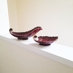 May include: Two red and gold leaf-shaped serving bowls. The larger bowl is curved and the smaller bowl is more rounded. Both bowls have a gold rim.