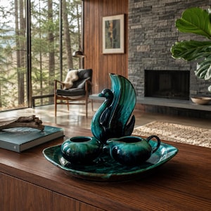 May include: A teal and dark green ceramic swan-shaped pitcher, a small bowl, and a creamer set arranged on a matching oval tray. The pieces feature a glossy, marbled glaze with deep blue and green tones, creating a cohesive set of serveware.