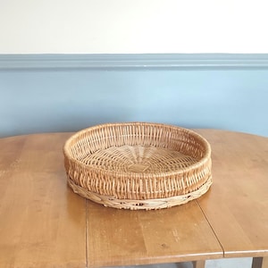 May include: A round, woven wicker basket with a natural brown color. The basket is sitting on a wooden table.