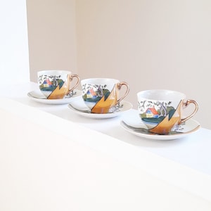 May include: Three white teacups and saucers with a landscape design. The cups have gold-coloured handles and a design of a house, trees, and a mountain. The saucers have a matching design. The cups and saucers are arranged on a white surface.