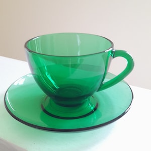 May include: A transparent emerald green teacup and saucer set. The cup has a rounded bowl with a curved handle. The saucer is circular with a slightly raised edge. The set is made of glass and has a simple, elegant design.