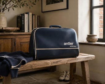 Pode incluir: Uma mala de desporto retangular azul-marinho com um acabamento liso e uma pega superior. A mala apresenta um detalhe de debrum horizontal de cor clara e o texto branco "antler" seguido de "SPORTBAG" impresso na parte frontal.