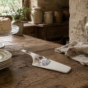 May include: A white ceramic cake server featuring a colourful floral pattern with pink, blue, and green details on the handle and blade. The server has a flat, triangular shape with a smooth finish and a metallic gold trim along the outer edge.