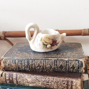 May include: A white ceramic swan trinket with a floral design featuring yellow and red roses. The swan is sitting on a stack of old books.