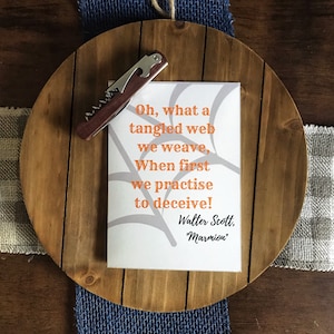 May include: A wooden wine serving tray with a canvas print featuring a quote by Walter Scott. A corkscrew rests on the tray. The quote reads, "Oh, what a tangled web we weave, When first we practise to deceive!"