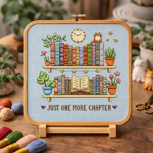 May include: A cross-stitch embroidery featuring a bookshelf design with colorful books, plants, and a cat. The text "JUST ONE MORE CHAPTER" is displayed below the shelves. The embroidery is set in a wooden frame.