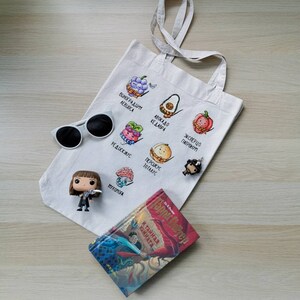 May include: A white tote bag with embroidered fruit illustrations and text in a foreign language. The bag is next to a pair of white sunglasses, a Harry Potter book, and a figurine. The book cover features a wizard and dragon.