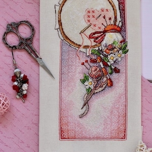 May include: A cross-stitch design featuring a wooden embroidery hoop with a heart-shaped pattern, needles, and red thread. The image includes a pair of silver scissors with a decorative charm, a pink pincushion, and floral accents. The background is a pink fabric.