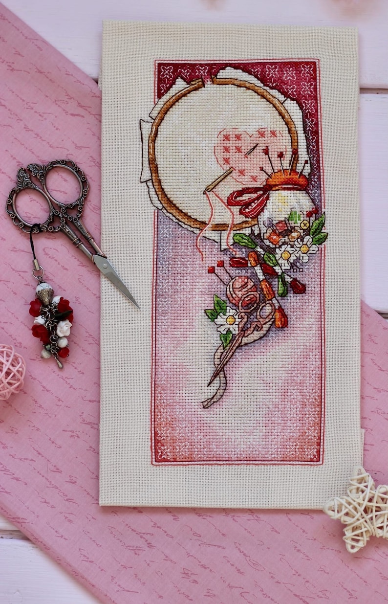 May include: A cross-stitch design featuring a wooden embroidery hoop with a heart-shaped pattern, needles, and red thread. The image includes a pair of silver scissors with a decorative charm, a pink pincushion, and floral accents. The background is a pink fabric.