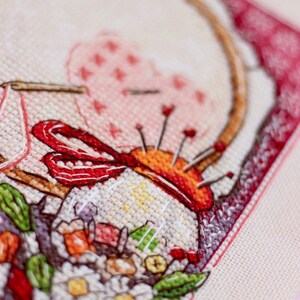 May include: Close-up of a cross-stitch embroidery featuring a needle holder with pins, a red ribbon, and a floral arrangement. The embroidery is framed with a red border and set against a light beige fabric.