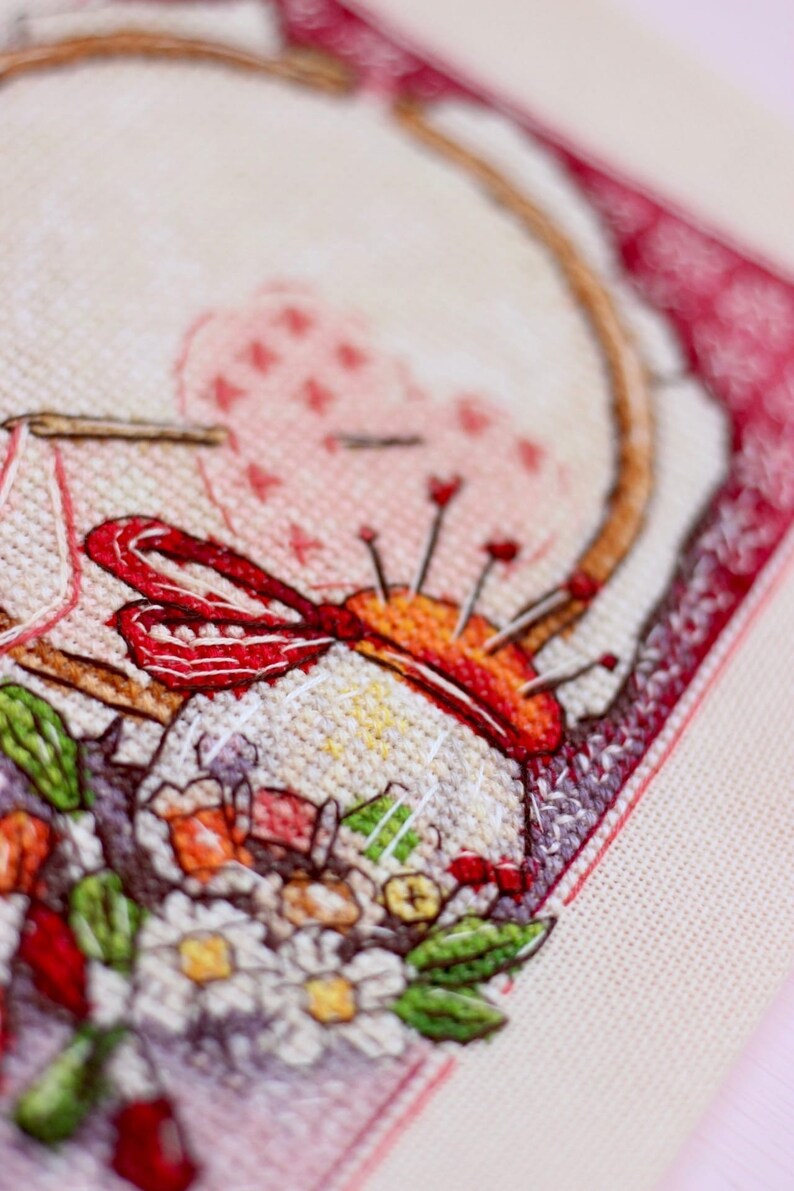 May include: Close-up of a cross-stitch embroidery featuring a needle holder with pins, a red ribbon, and a floral arrangement. The embroidery is framed with a red border and set against a light beige fabric.