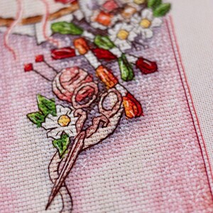 May include: Close-up of a cross-stitch design featuring a pink yarn ball, scissors, needles, and a bouquet of flowers. The design is on a pink fabric background, with visible cross-stitch details. The image evokes a crafting or needlework theme.