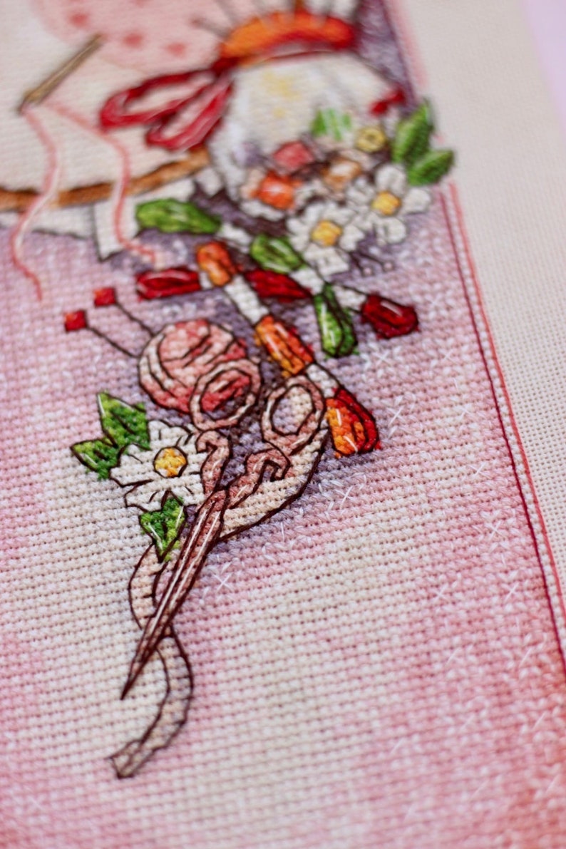 May include: Close-up of a cross-stitch design featuring a pink yarn ball, scissors, needles, and a bouquet of flowers. The design is on a pink fabric background, with visible cross-stitch details. The image evokes a crafting or needlework theme.
