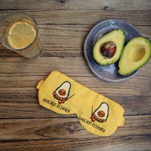 May include: Yellow sleep mask with embroidered avocado design and the text "AVOCADO KEDAVRA". The mask features a cartoon avocado with glasses and a scarf. A glass of water with a lemon slice and a plate with avocado halves are also visible.