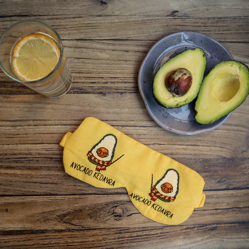 May include: Yellow sleep mask with embroidered avocado design and the text "AVOCADO KEDAVRA". The mask features a cartoon avocado with glasses and a scarf. A glass of water with a lemon slice and a plate with avocado halves are also visible.