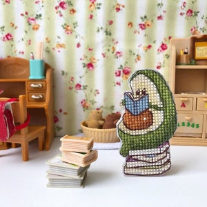 May include: A green avocado-shaped plush toy is sitting on a stack of books. The avocado is holding a blue book and has a smiling face. The avocado is sitting on a white surface.
